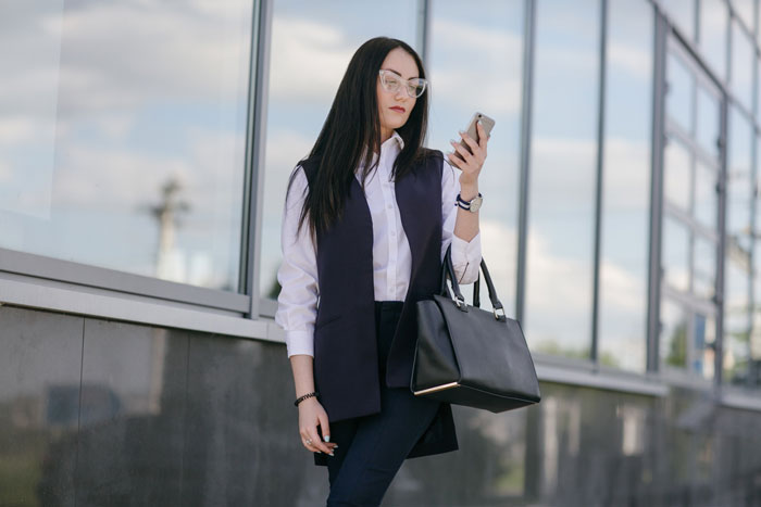 Young woman in professional attire looking at smartphone outside office, reflecting cheater lady and animal-lover coworker themes.