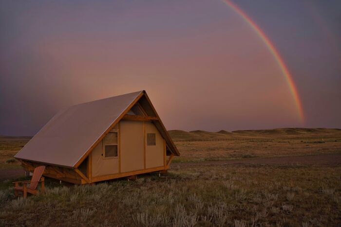 A rainbow arcs over a tan glamping tent in a vast, grassy field at dusk, offering a gentle reset.