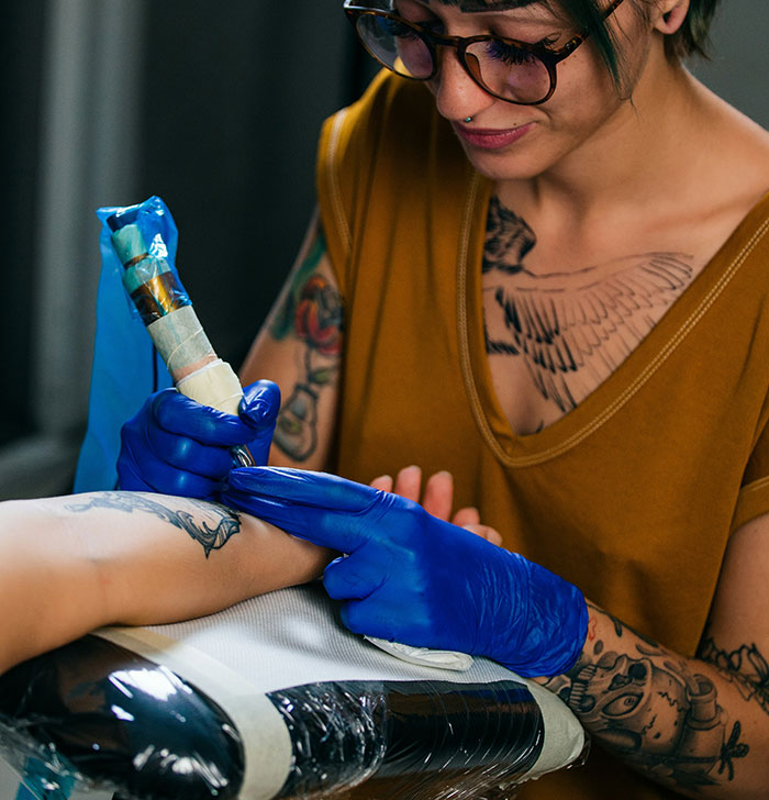 Tattoo artist with glasses and gloves creating detailed ink work on a client’s arm, showcasing tattoo culture and skill.