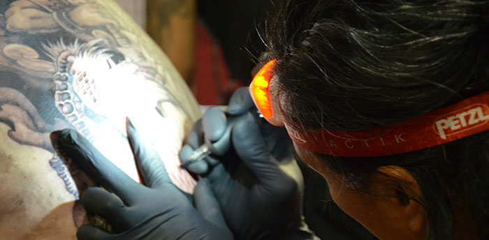 Tattoo artist wearing a headlamp tattooing intricate Chinese and Japanese characters on a person's shoulder in a dimly lit room.