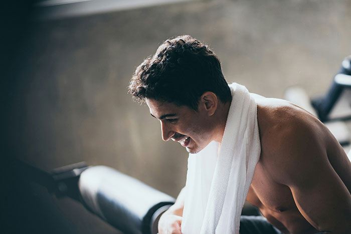 Young man with towel on shoulders at gym, smiling while resting after workout, highlighting tattoos and body art trends
