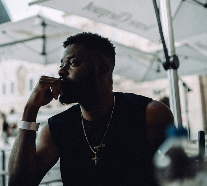Man with beard sitting thoughtfully under umbrella, highlighting tattoos and people amused by Chinese and Japanese tattoos.