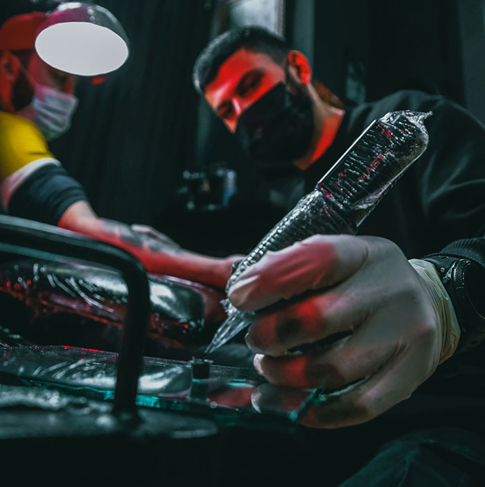 Tattoo artist wearing gloves working on a client’s arm with Chinese and Japanese tattoo designs in a dim studio.