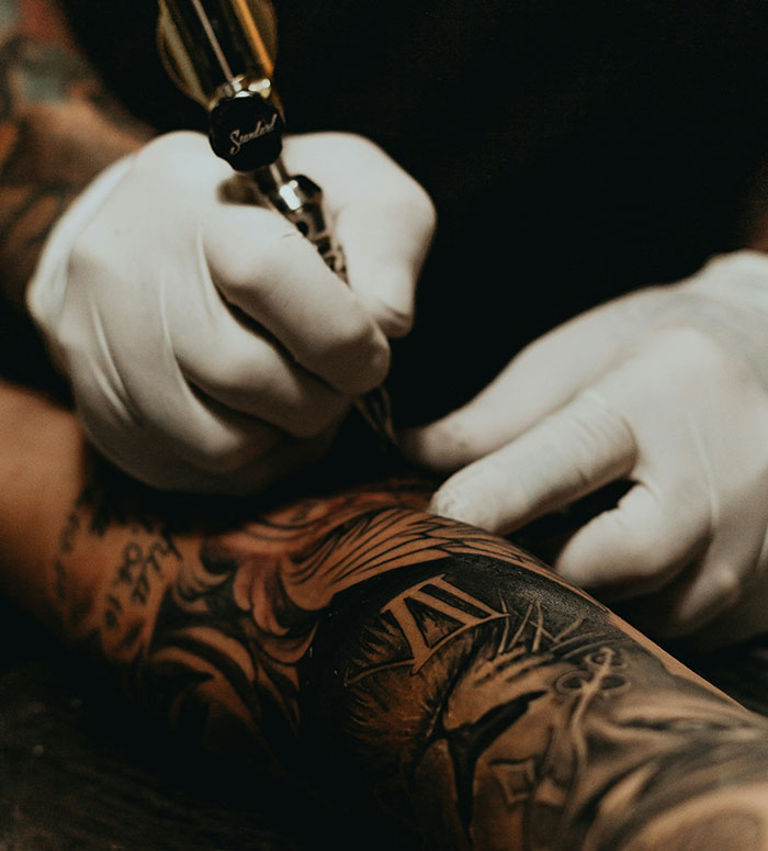 Close-up of a tattoo artist wearing gloves creating detailed ink work on a heavily tattooed arm with Chinese and Japanese characters.