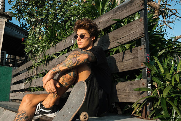 Young man with detailed tattoos on his arms and legs sitting outdoors with a skateboard, showcasing unique tattoo designs.