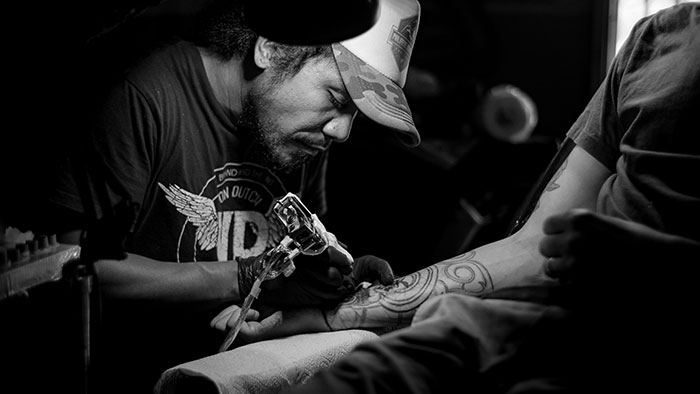 Tattoo artist in a cap creating a detailed tattoo on a client's arm, showcasing Chinese and Japanese tattoo art styles.