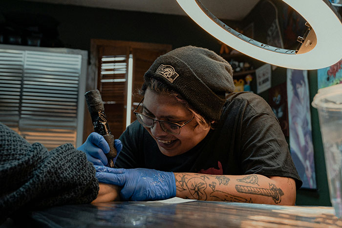 Tattoo artist with glasses and beanie smiling while working on a detailed tattoo in a cozy studio setting.