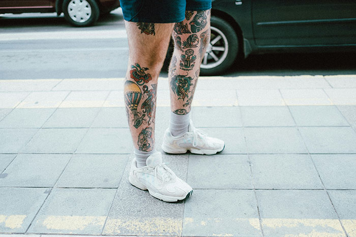 Legs with various colorful tattoos visible, including traditional and modern designs, on a city sidewalk near parked cars.