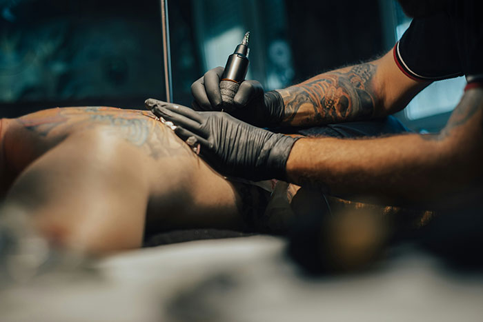 Tattoo artist with gloves using a machine to ink a client's back, highlighting tattoos involving Chinese and Japanese characters.