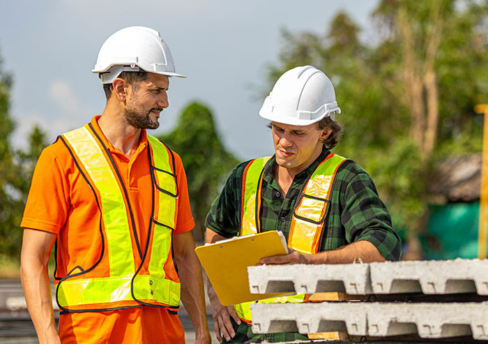 Two construction coworkers in safety vests and helmets reviewing plans beside stacked concrete slabs outdoors