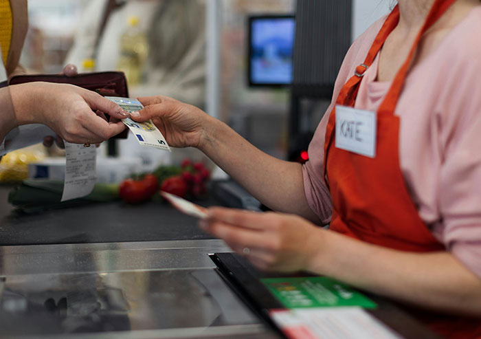 Cashier coworker wearing an apron named Kate accepting a five euro bill at a grocery store checkout counter.