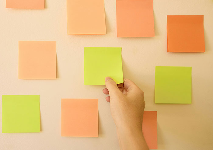 Hand placing blank green sticky note on wall among other orange and green notes, illustrating coworker mistakes concept.