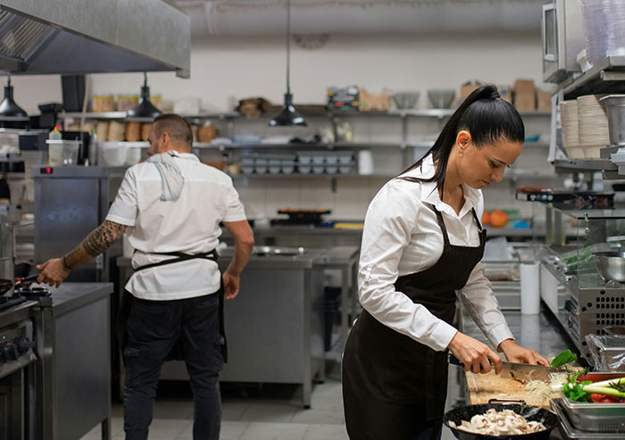 Two coworkers working in a busy commercial kitchen, with one chopping vegetables and the other cooking at the stove.