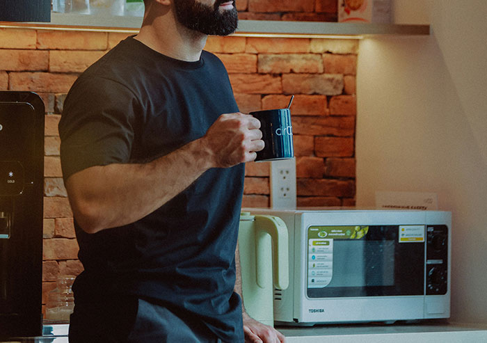 Man holding mug in a kitchen area with a microwave and brick wall, illustrating coworkers who took stupidity to new heights.