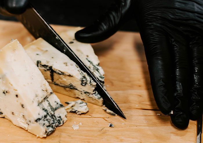Hand in black glove cutting blue cheese with a knife on a wooden board, showing coworkers' unexpected actions.