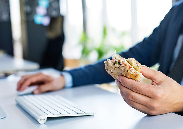 A coworker eating a sandwich with one hand while typing on a keyboard in an office setting showing coworker behavior.