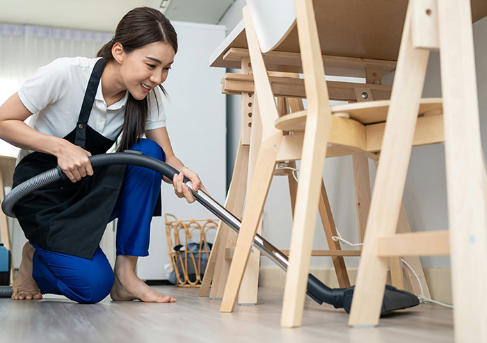 Young woman coworker using vacuum cleaner under wooden chairs, showcasing unexpected office cleaning moments in coworker behavior.