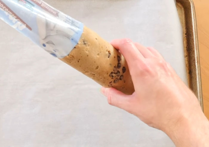 Hand holding cookie dough tube over baking tray, illustrating coworker mistakes involving cooking in office kitchens.