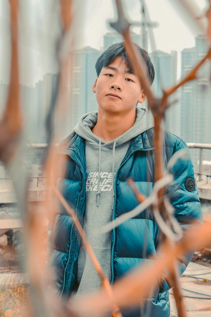 Young man wearing a blue jacket and hoodie, posing outdoors in an urban setting, displaying intelligence and confidence.