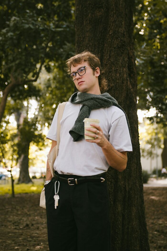 Young man leaning on tree holding a drink, wearing glasses and scarf, showcasing intelligence in a casual outdoor setting.