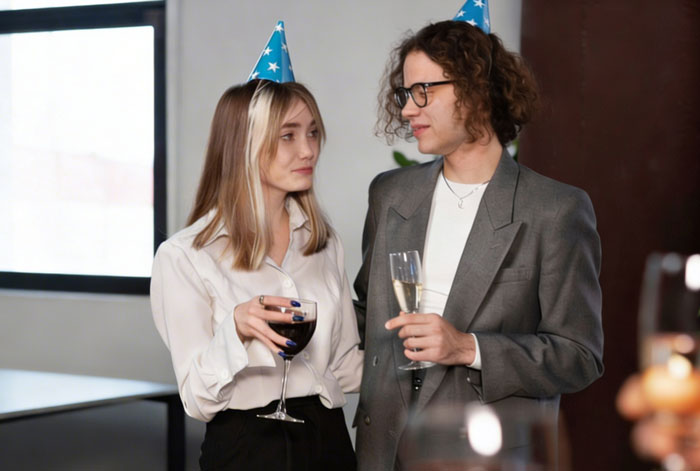 Two employees wearing party hats holding drinks at a company celebration with balloons and lunch event.