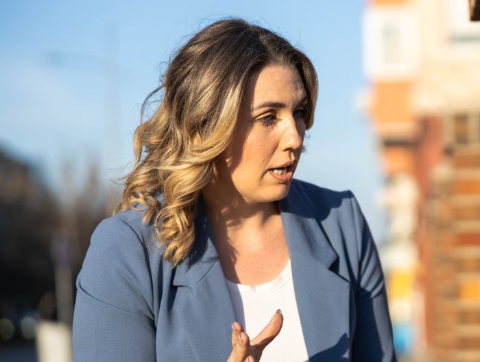 A blonde woman in a blue blazer, speaking and gesturing outdoors about trying to transition her son into vegan.