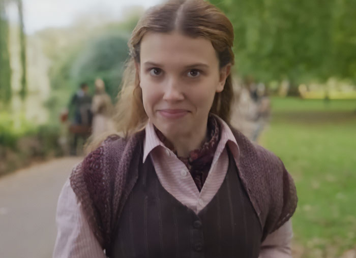 Millie Bobby Brown as Enola Holmes 3, smiling in a park with trees and blurred figures in the background.