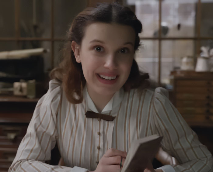 Millie Bobby Brown as Enola Holmes 3, smiling brightly in a striped shirt, holding a small book.