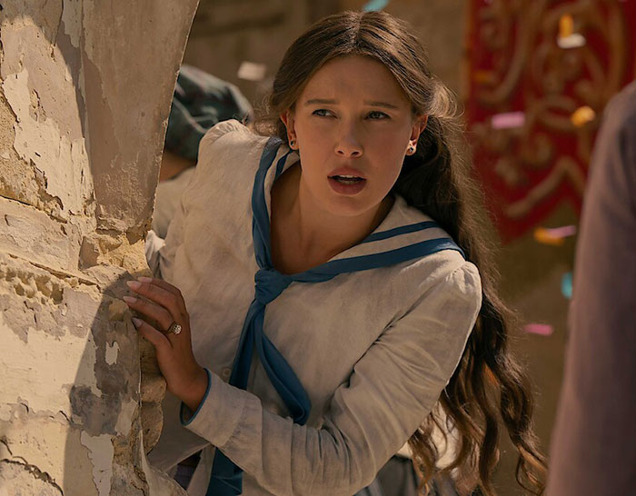 Millie Bobby Brown as Enola Holmes, looking concerned and peering from behind a stone wall. First-look photos.