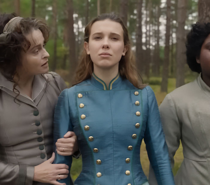 Millie Bobby Brown as Enola Holmes, in a blue military-style jacket, being held by another woman in a forest setting.