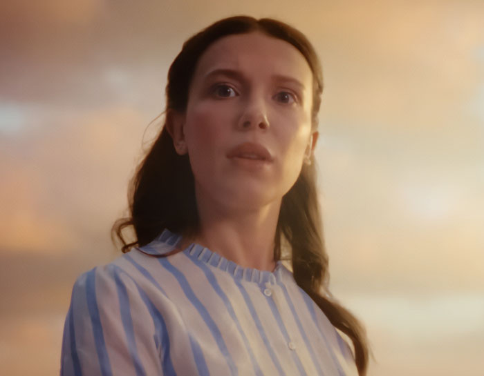 Millie Bobby Brown as Enola Holmes, wearing a striped shirt, looking up with a thoughtful expression for Enola Holmes 3.