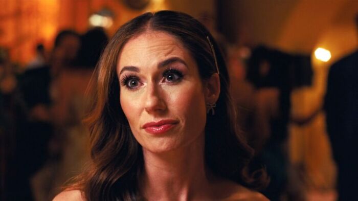 Sydney Sweeney from Euphoria with a slight smirk, hair down, and a gold clip, amidst a blurred background.