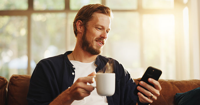 Man holding a smartphone and drinking coffee while sitting on a couch, symbolizing woman running Tinder scams for jailed fianc&eacute;.