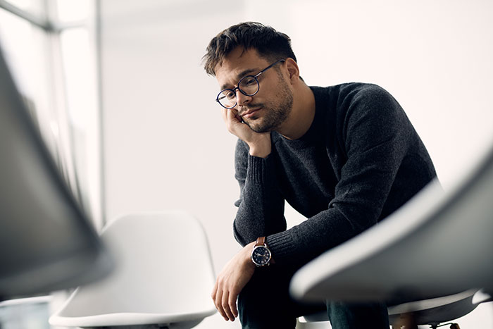 Young man wearing glasses and dark sweater sitting thoughtfully in a chair representing a Tinder scam victim.