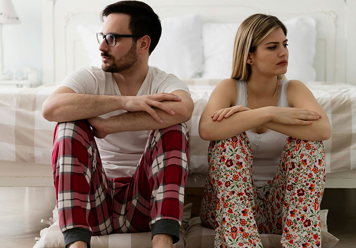Couple sitting apart on bedroom floor in pajamas, looking upset, reflecting family vacation and stepson disagreement.