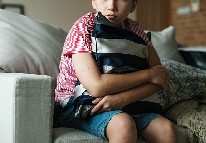 Young boy hugging pillow on couch, appearing upset as woman pays for family vacation without him.