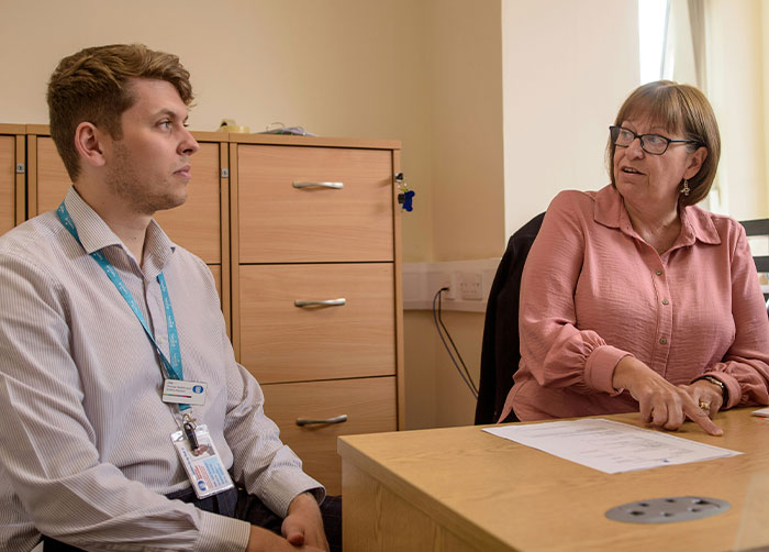 Two people in an office. A man listens as a woman gestures to a document, planning perfect revenge on a horrible boss.