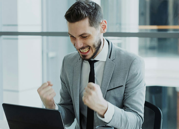 A man in a suit clenches his fists in triumph, smiling wide at a laptop. The perfect revenge for a horrible boss.