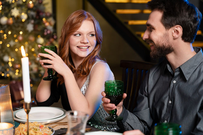 Man plays expert at dinner party, holding green glass and smiling at real expert who questions him.