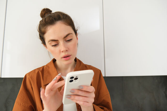 Young woman in a brown jacket looking surprised while checking DNA test results on her smartphone in a modern kitchen.