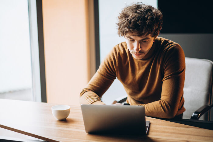 Young man researching DNA tests and ancestry results on laptop while seated at a wooden desk near a window with coffee cup.