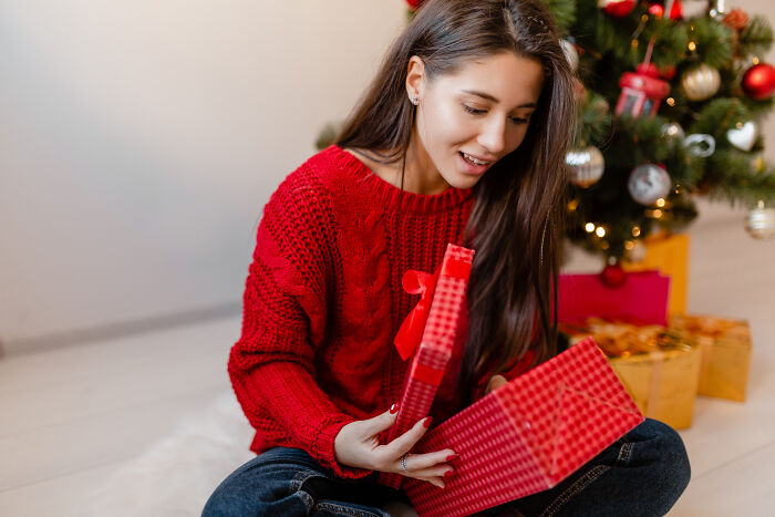 Young woman in a red sweater opening a gift box near a decorated Christmas tree, symbolizing DNA test surprises.