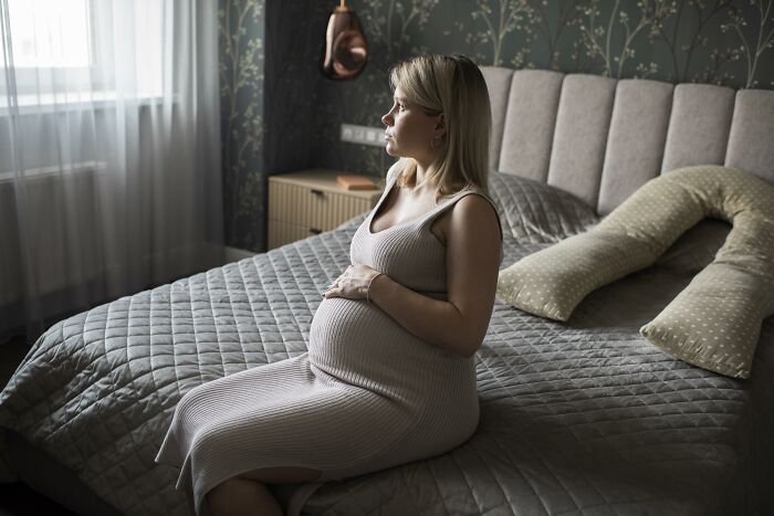Pregnant woman sitting on bed, looking thoughtfully out window, reflecting on unexpected DNA test results.
