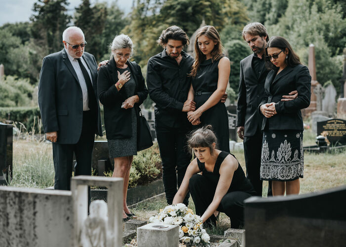 A group of people in black attire at a gravesite, one woman placing flowers. They look sad and contemplative, like finding out they were cheated on.