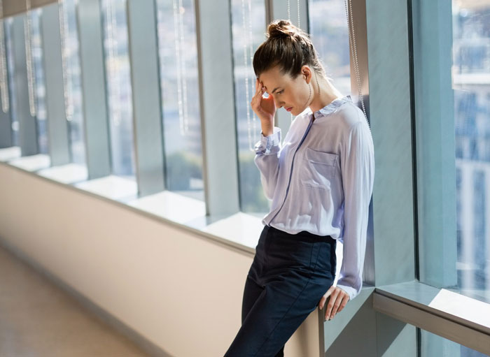 Woman in an office, leaning against a window, looking stressed. Highlights challenges for a woman with a feeding tube.