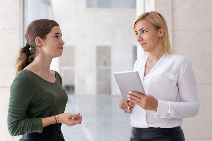 Two women in an office hallway; one speaks while the other, holding a tablet, looks skeptically. This represents challenges for a woman relying on a feeding tube.