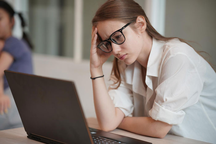 A woman with a feeding tube, wearing glasses and a white shirt, looks frustrated while working on her laptop.