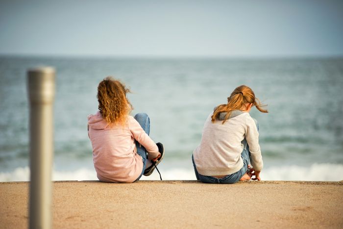 Two girls sit on a concrete ledge, backs to the camera, looking at the ocean. A best friend skips wedding.