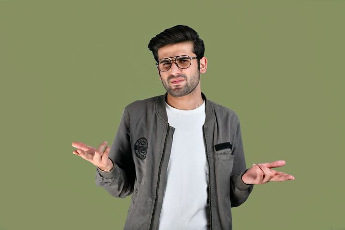 Man in glasses and casual jacket shrugging with confused expression against a plain green background, reflecting wild times employees quitting.
