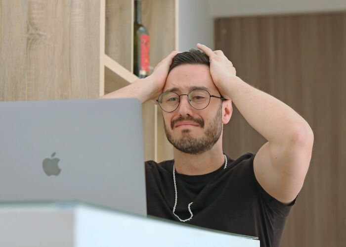 Man wearing glasses, holding his head in frustration while looking at a laptop after seeing an accidental message sent out.